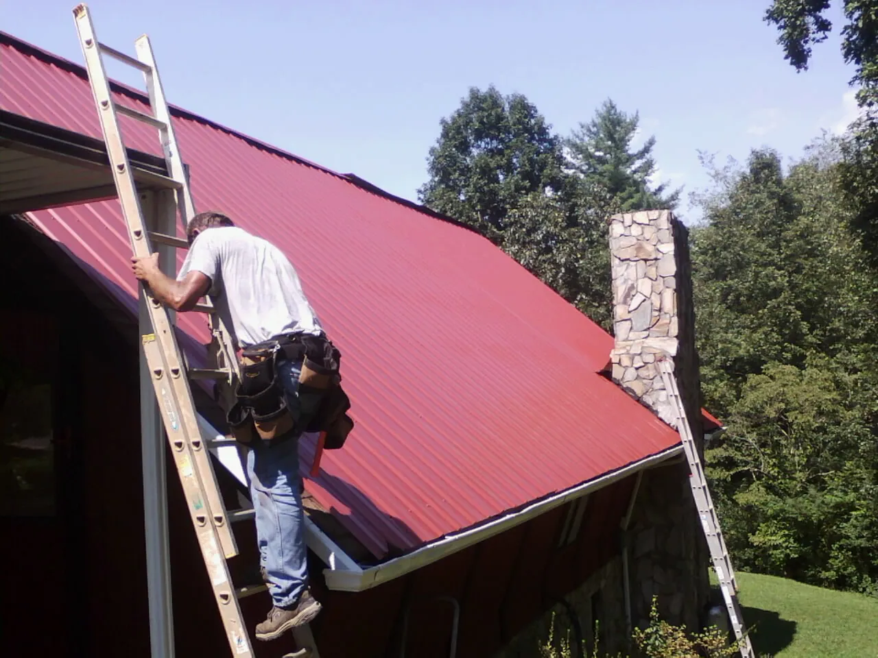 Roofer on a ladder beside a red metal roof and stone chimney — Soffit & Fascia Repair by Roofing Brothers in Guys Mills